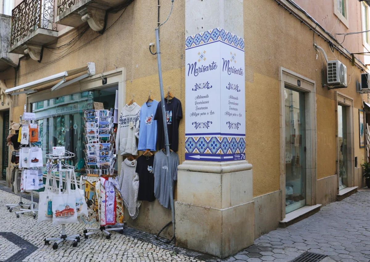 A esquina da loja, com camisolas, sacos de pano e postais expostos à porta, e o painel de azulejo com o nome Marisarte
